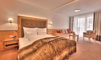A cozy bedroom with a large bed and bright walls. There is a seating area with a sofa and plenty of daylight from the windows.
