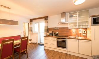 A modern kitchen with white furniture and wooden flooring. There is a dining area with red chairs and a large window for daylight.
