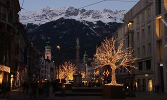 A festively lit street with glittering trees. In the background, snow-covered mountains and historic buildings can be seen.