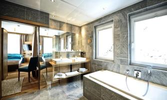 A modern bathroom with a bright stone floor, bathtub, and two sinks. In the background, a cozy bedroom with blue beds is visible.