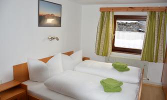 A cozy bedroom with two single beds, white bedspreads, and green towels. The window offers a view of the snowy landscape outside.