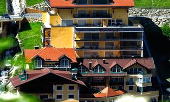 A picturesque hotel building with several floors, surrounded by green mountains. The hotel offers wellness, an indoor pool, and a sauna.