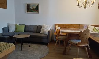 A cozy living space with a gray sofa, a round table, and a rustic dining table. The walls are painted light, and there is a wall lantern as well as a picturesque painting.