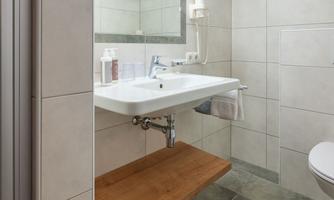 A modern bathroom with white tiles and a large mirror. There is a sink with a wooden shelf and a toilet.