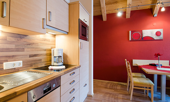 Modern kitchen with wooden furniture and functional appliances. In the background, there is a dining area with a red accent wall and a decorative table.
