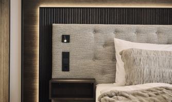 A modern bed with a padded headboard and soft lighting. Next to the bed is a small nightstand with a power outlet.