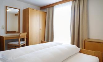 A simple bedroom with two beds, a table, and a chair. The room is bright and has large windows with curtains.