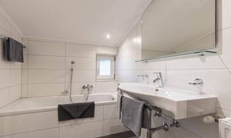 A modern bathroom with a bathtub and a sink. The walls are light tiled and there is a window that lets in natural light.