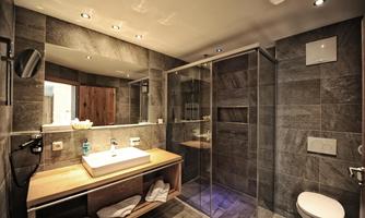 A modern bathroom with a shower and a sink on a wooden shelf. The walls are covered with gray tiles and there is indirect lighting.