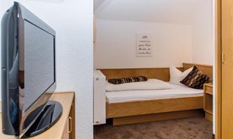 A cozy room with a wooden bed and a television on a nightstand. The walls are light and there are cuddly pillows on the bed.
