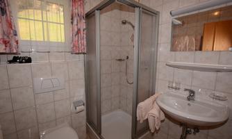 A small bathroom with a shower cubicle, sink, and toilet. It has light tiles and a window with curtains.