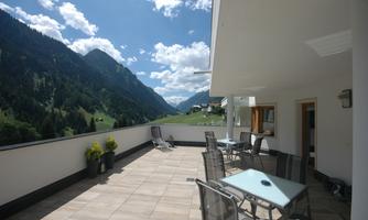 A sunny terrace with tables and chairs, surrounded by green mountains. The sky is blue with some clouds.
