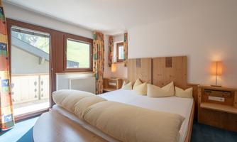 A cozy bedroom with a large bed and bright pillows. There are windows with a view and a warmly lit bedside lamp.