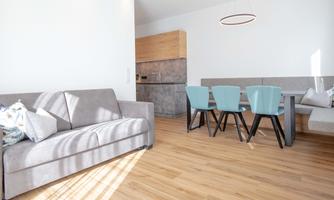 A bright and modern living room with a gray sofa and a dining table with blue chairs. The wooden floors and the open kitchen give the space an inviting atmosphere.