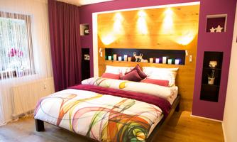 A modern bedroom with a large bed and colorful cushions. The walls are designed in purple and wood colors, with soft lighting in the background.