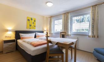 A cozy bedroom with a double bed and a table. Bright walls and large windows create a friendly atmosphere.