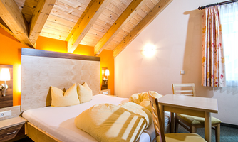 A cozy room with a wooden ceiling, a double bed, and a small table. The walls are painted in warm colors, and the room is well lit.