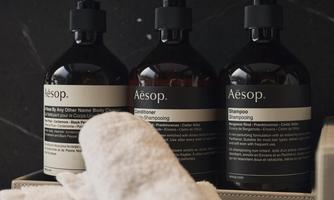 Three bottles of Aesop care products are standing on a table, surrounded by white towels. The bottles have black pumps and a stylish design.