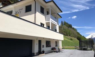 A modern house in a picturesque landscape, surrounded by green hills and a clear blue sky. The facade is bright and friendly, featuring a parking space and outdoor seating.