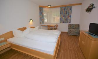 A cozy hotel room with two beds, a table, and an armchair. The room has a large window with curtains and wooden flooring.