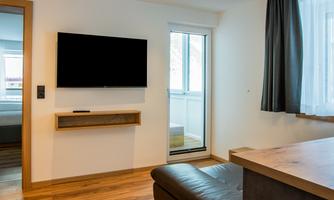 A modern room with a flat-screen TV on the wall and a large window. The floor is made of wood and there is a cozy sofa and a table.
