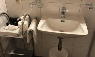 A modern bathroom with a sink, a soap dispenser, and a wastebasket. Towels and toiletries are neatly arranged on a shelf.