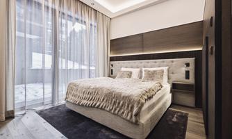 A modern bedroom with a large bed and soft blankets. Large windows let in light and offer a view of the winter landscape outside.