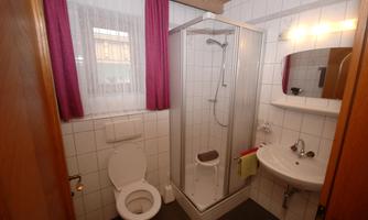A small bathroom with a shower, toilet, and sink. The walls are tiled, and there are windows with curtains.