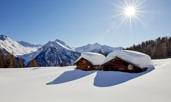 A winter landscape with two old wooden cabins covered in snow. In the background, impressive mountains rise beneath a clear blue sky.
