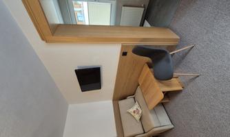 A small room with a cozy sofa, a table, and a television on the wall. In the background, there is a window area overlooking another room.