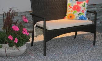 A cozy rattan bench with a colorful cushion sits on a gravel floor. Next to the bench, pink flowers bloom in a pot.