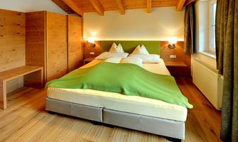 A cozy bedroom with a large bed and a green blanket. The walls are made of wood, and there are large windows that let in plenty of light.