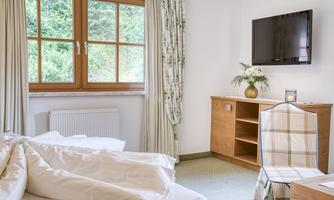 A cozy bedroom with a window that lets in daylight. There is a TV, a wooden wardrobe, and a chair next to a table.