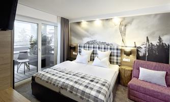 A modern hotel room with a double bed and a cozy armchair. Large windows offer a view of a balcony and a mountain landscape.