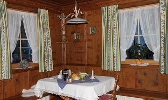 A cozy dining area with wooden walls and a table adorned with a tablecloth. Curtains with a floral pattern hang in front of the windows, and there are some decorative elements visible.