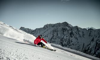 A skier in red is skiing through fresh snow in front of an impressive mountain landscape. The sky is clear and the mountain peaks are covered in snow.
