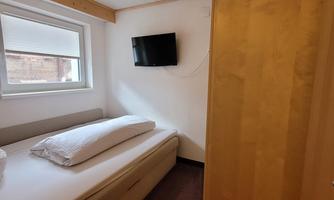 A simple bedroom with a single bed and a television on the wall. There is a small table and a window that provides daylight.