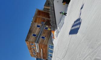 A modern building on a snowy slope under a bright blue sky. Winter sports enthusiasts can be seen nearby.