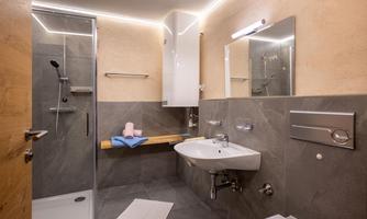 A modern bathroom with a shower, mirror, and a sink. The walls are elegantly designed, and there are towels on a wooden shelf.