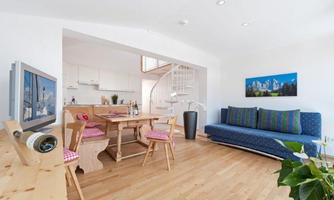 A modern living room with wooden flooring and a cozy sofa. In the background, an open kitchen and a dining table with chairs can be seen.