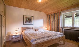 A cozy bedroom with a wooden double bed and a warm wooden ceiling. There are two nightstands with lamps and a colorful curtain at the window.