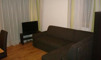 A simply furnished living space with a dark gray sofa and a television. Large windows with curtains let in plenty of light.