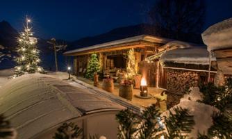 A cozy wooden house in the snow, surrounded by festively decorated fir trees. The atmosphere is calm and inviting at dusk.