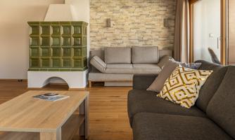 A modern apartment with a stylish gray sofa and a cozy wooden floor. In the background, there is a green tiled stove and a large window view.