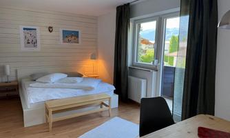 A modern bedroom with a large bed and bright colors. A window leads to a balcony with a view of the outdoors.