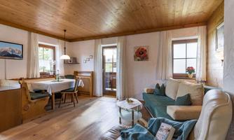 A cozy living room with wooden walls and a wooden floor. There is a sofa, a dining table, and windows with a view outside.