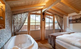 A cozy bedroom with wooden beams and large windows. A bathtub is next to the bed, and the room is tastefully decorated.