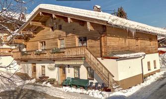A traditional wooden house in the snow, surrounded by a wintry landscape. The roof is covered with snow, and there is a clear blue sky in the background.