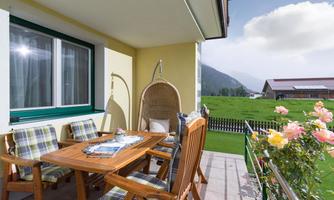 A cozy balcony with wooden chairs and a dining table. In the background, green meadows and mountains can be seen.
