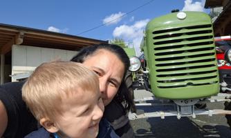 A woman smiles with a small boy in front of a green tractor. The sky is blue with a few clouds.